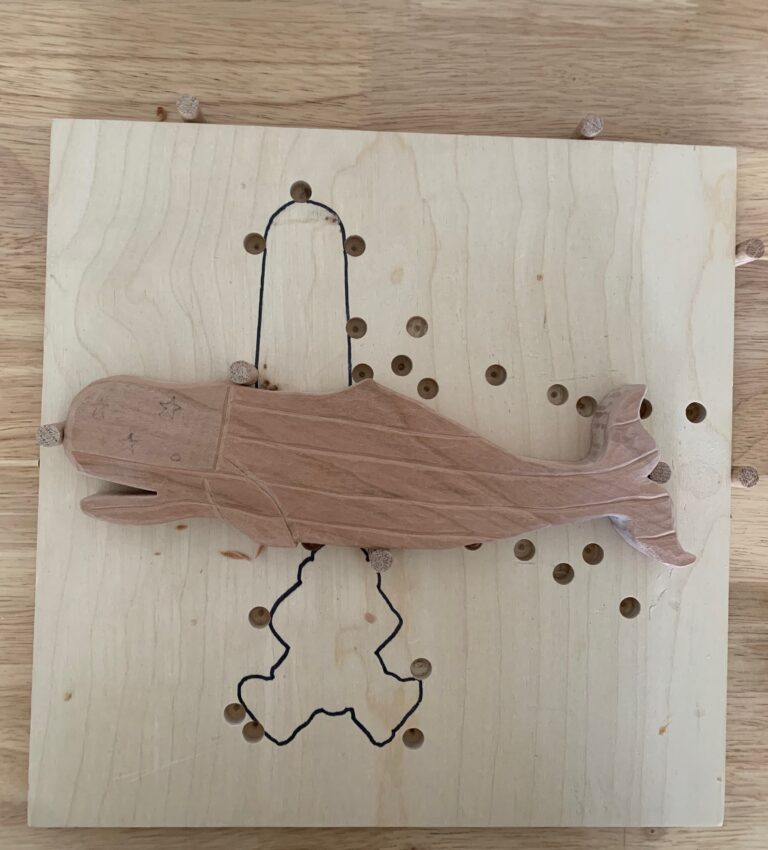 Wooden fish carving held between dowel pegs on a homemade plywood holding board.