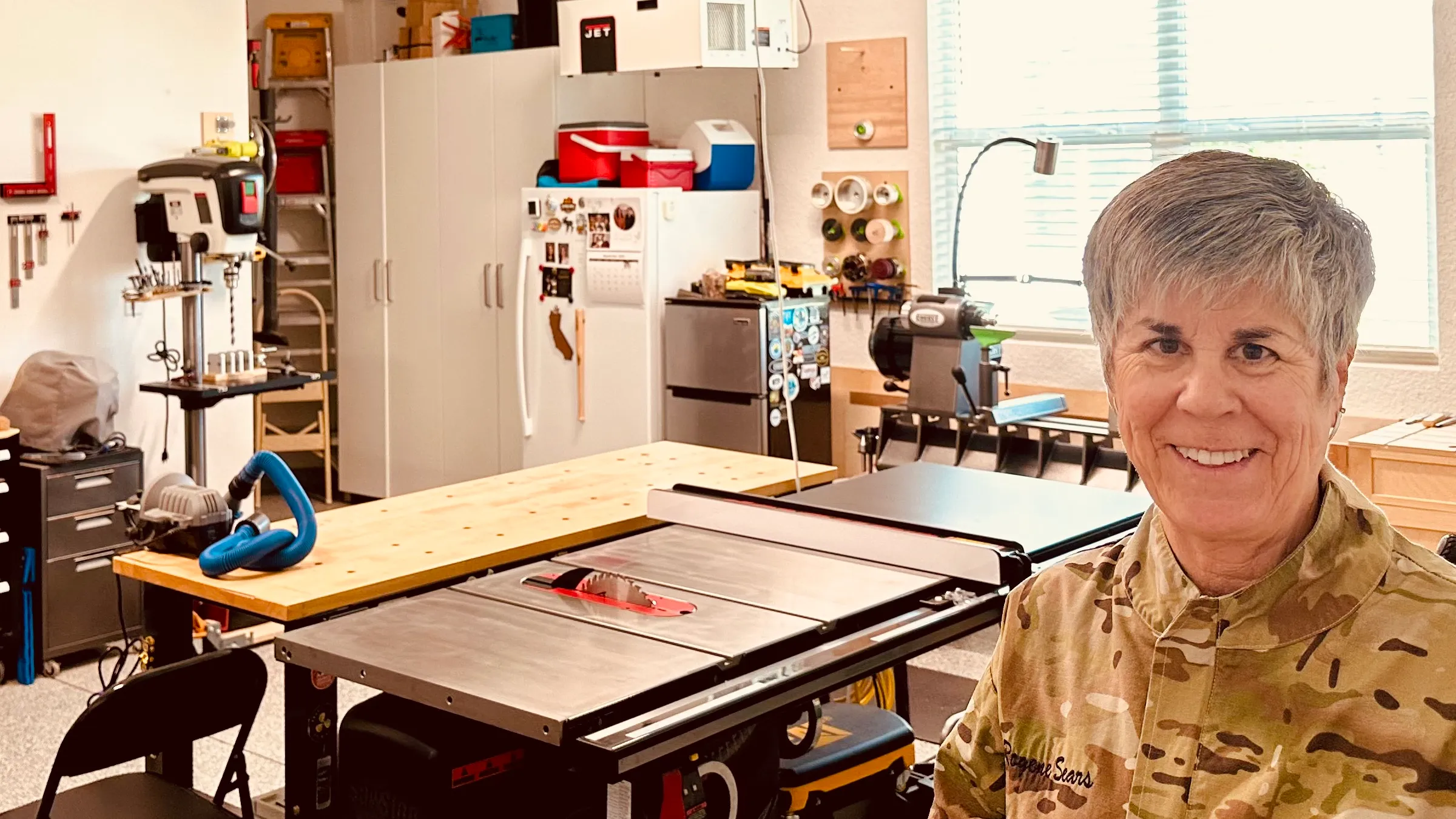 Rogene Sears smiling in the Chipin’ Wood workshop, with a table saw and lathe nearby.
