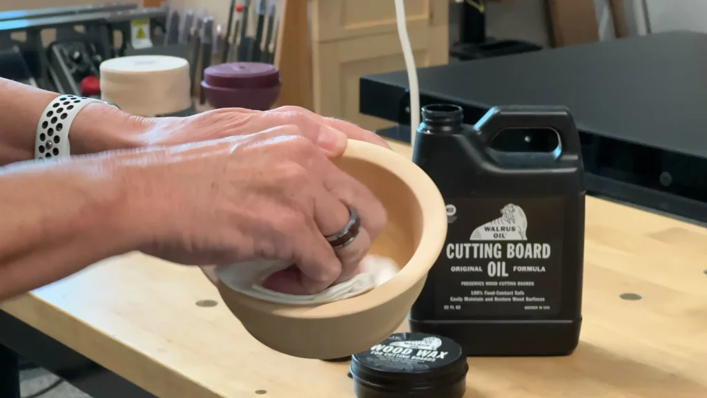 Hands applying oil inside a wooden bowl beside Walrus Oil Cutting Board Oil and Wood Wax.