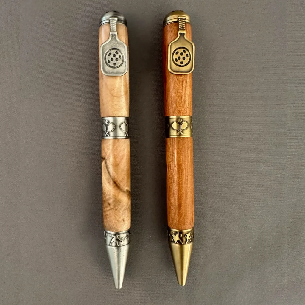 Two wooden pickleball pens with paddle clips, shown in antique pewter and antique brass finishes.