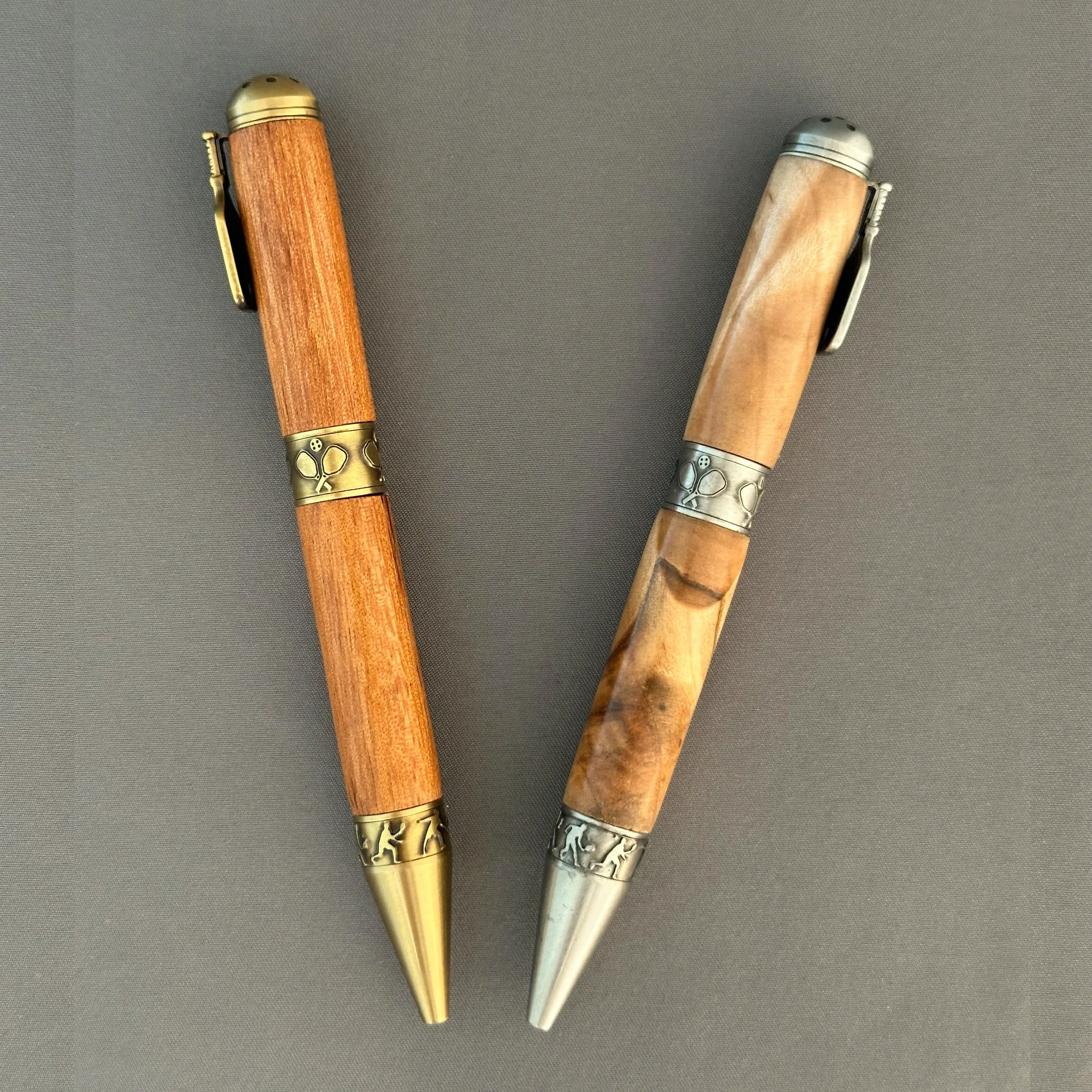 Two wooden pickleball pens arranged in a V, showing their contrasting grain and antique brass and pewter hardware.