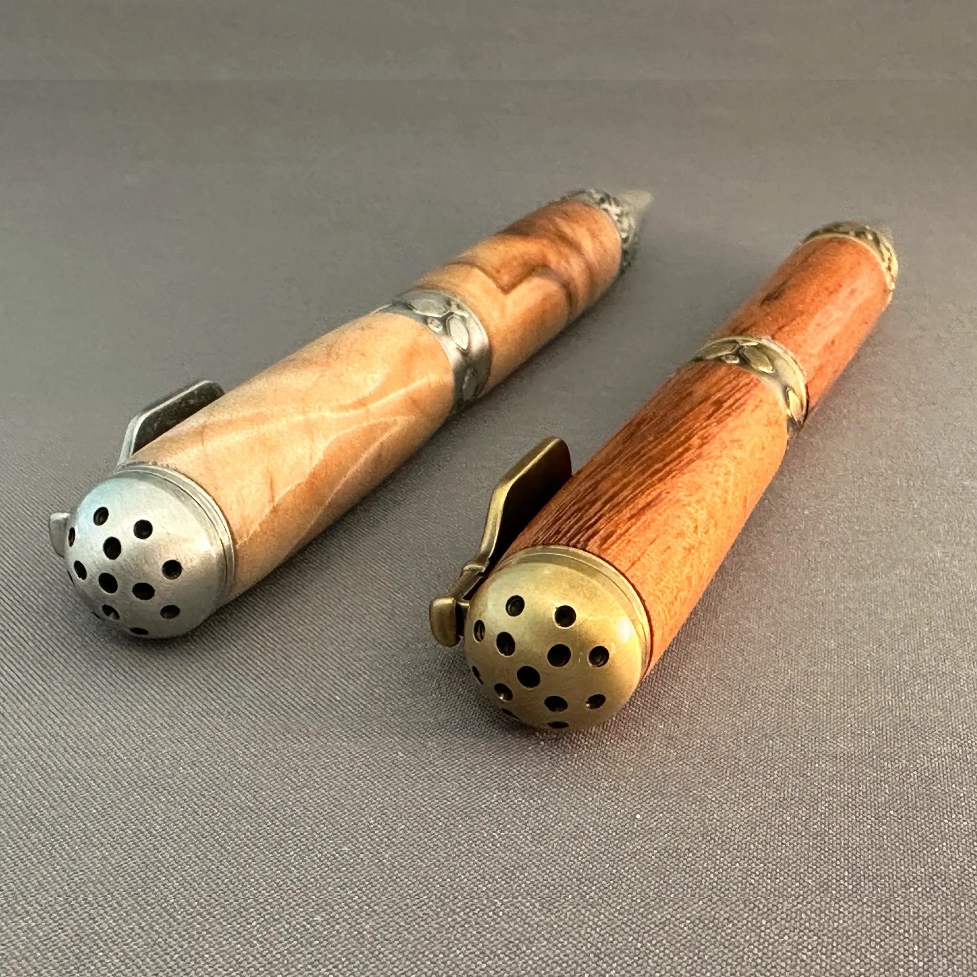 Close-up of perforated pickleball-shaped end caps in antique pewter and antique brass on two wooden pens.