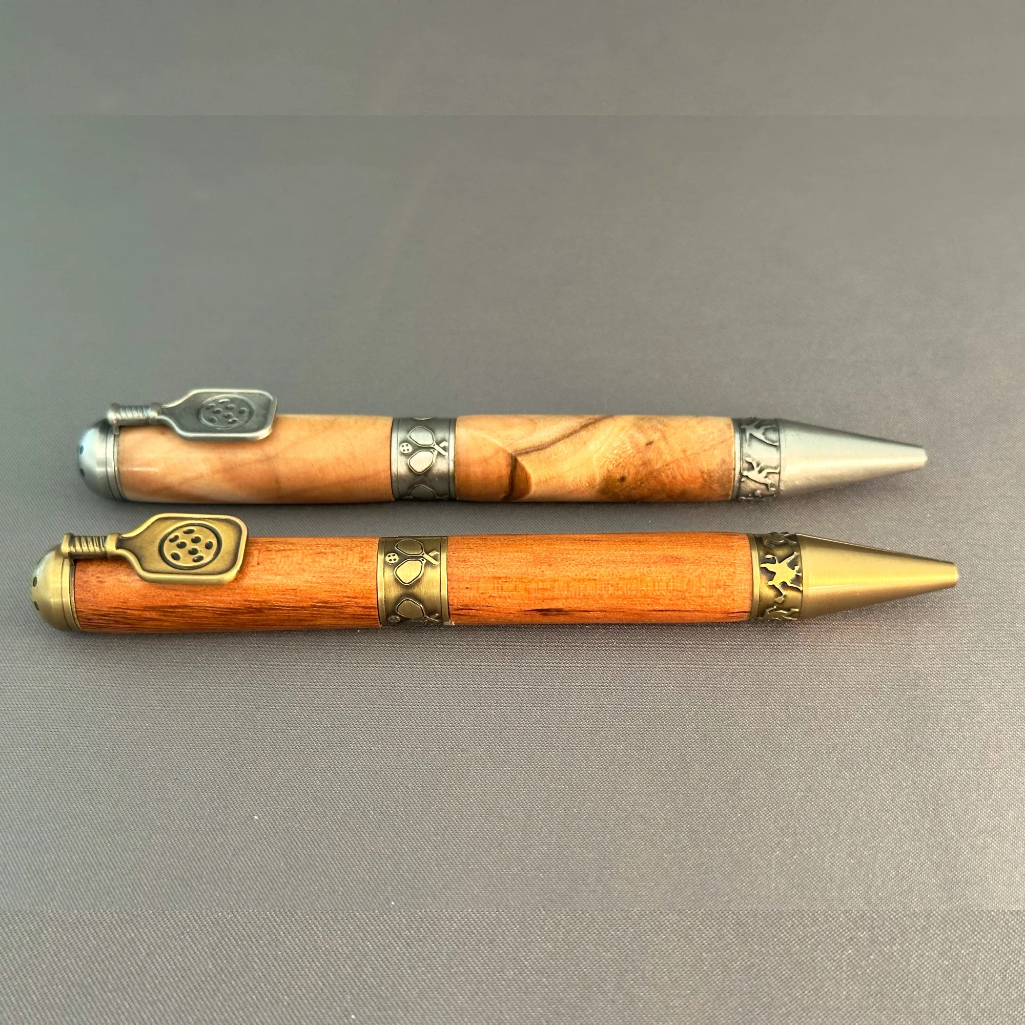 Two wooden pickleball pens displayed horizontally, with antique brass above antique pewter and both paddle clips visible.