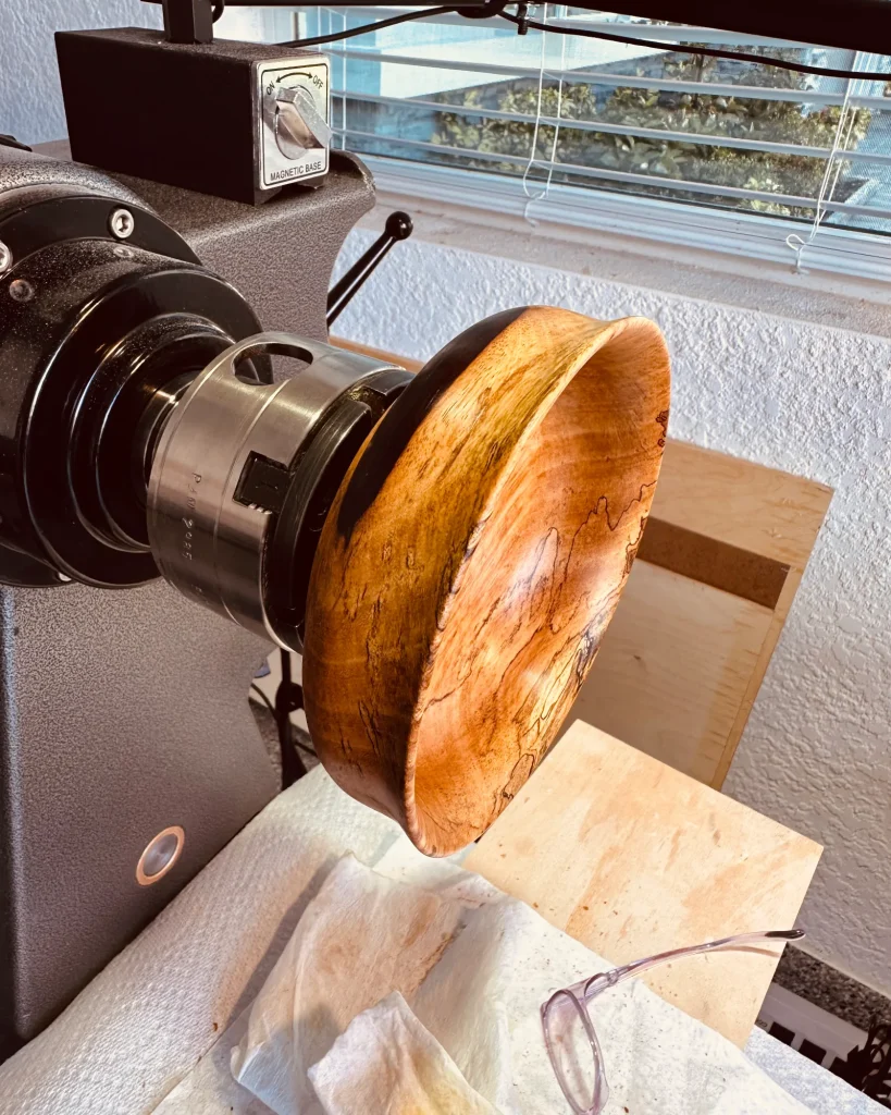 Wooden bowl mounted on a lathe, showing its curved rim, warm golden color, and dark spalting lines.